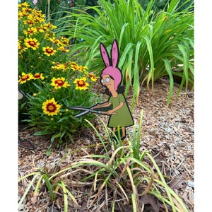 Puede incluir: Un adorno de jard&iacute;n plano con un personaje de dibujos animados de pelo oscuro, que lleva un gorro rosa con orejas de conejo y un vestido verde, sosteniendo unas tijeras de jard&iacute;n de metal. El personaje est&aacute; situado entre follaje y flores amarillas.