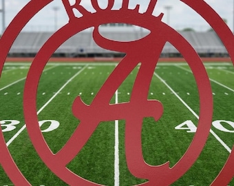 Op de afbeelding: Rood metalen bord met de woorden "ROLL TIDE" en een grote letter "A" in een cirkel. Het bord hangt aan een ketting tegen een achtergrond van een voetbalveld. Een decoratief item voor fans.
