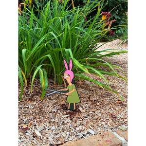 Puede incluir: Un adorno de jard&iacute;n plano con un personaje de dibujos animados que lleva un gorro rosa con orejas de conejo y un vestido verde, sosteniendo unas tijeras de podar plateadas. El personaje est&aacute; colocado sobre un lecho de mantillo de madera.