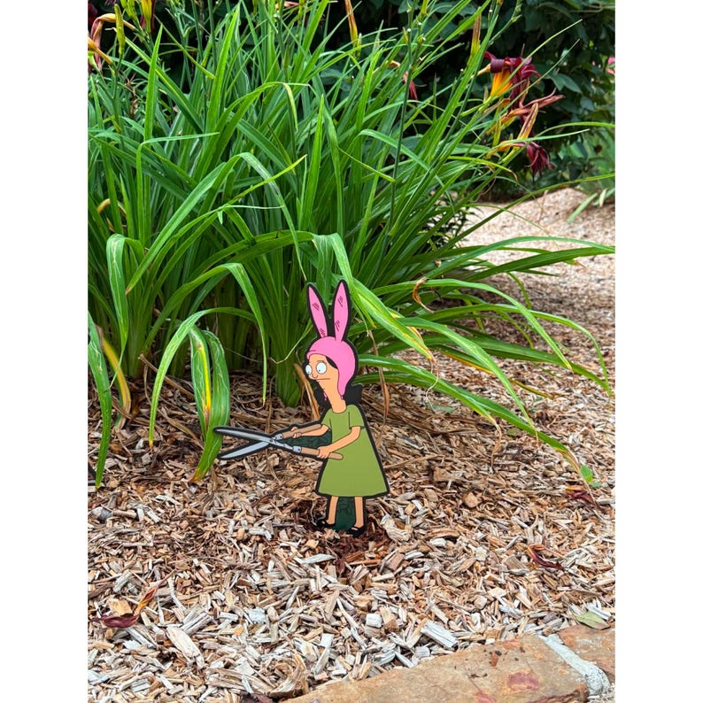 Puede incluir: Un adorno de jard&iacute;n plano con un personaje de dibujos animados que lleva un gorro rosa con orejas de conejo y un vestido verde, sosteniendo unas tijeras de podar plateadas. El personaje est&aacute; colocado sobre un lecho de mantillo de madera.