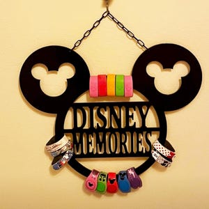 May include: Black Mickey Mouse-shaped sign with the words "DISNEY MEMORIES" cut out. The sign holds colorful wristbands and is suspended by a black chain. The sign has two large Mickey Mouse ears with the iconic Mickey Mouse head shape.
