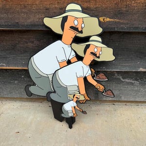 Puede incluir: Un recorte de madera en capas que muestra tres figuras de dibujos animados id&eacute;nticas con bigote, que llevan un sombrero beige de ala ancha, una camiseta blanca y pantalones grises. Cada figura est&aacute; arrodillada y sostiene una peque&ntilde;a paleta.