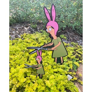 Puede incluir: Dos letreros de jard&iacute;n planos con forma de silueta que muestran un personaje de dibujos animados con cabello negro, un gorro rosa con orejas de conejo y un vestido verde. El personaje aparece en dos tama&ntilde;os, sosteniendo tijeras de podar.