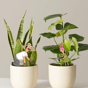 Puede incluir: Dos macetas de cer&aacute;mica color hueso que contienen una planta de serpiente y una monstera. Un personaje de dibujos animados con bigote y sombrero de paja se arrodilla en la planta de serpiente, mientras otro con orejas de conejo rosas poda la monstera con tijeras de metal.