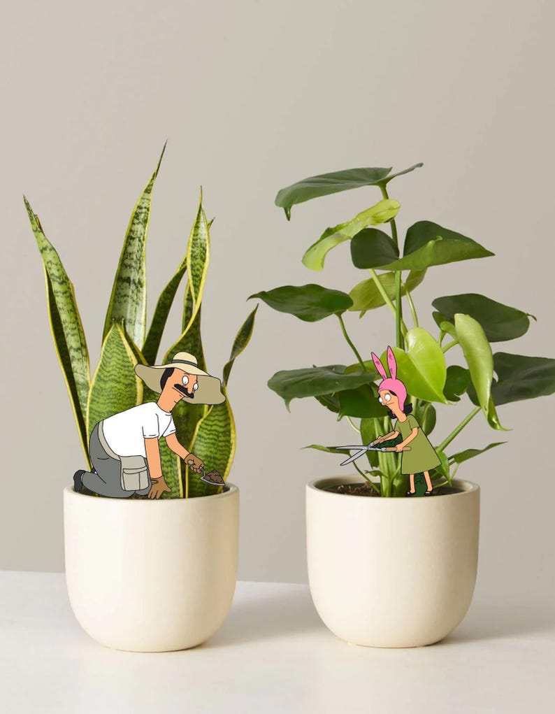 Puede incluir: Dos macetas de cer&aacute;mica color hueso que contienen una planta de serpiente y una monstera. Un personaje de dibujos animados con bigote y sombrero de paja se arrodilla en la planta de serpiente, mientras otro con orejas de conejo rosas poda la monstera con tijeras de metal.