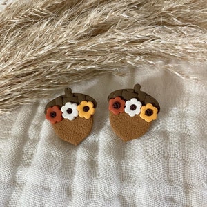 May include: Two acorn-shaped earrings. The earrings are brown with a dark brown cap and decorated with small, colourful flowers in orange, white, and yellow. They are set against a white, textured background.