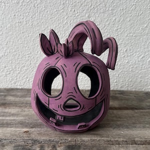 May include: A purple ceramic pumpkin lantern featuring a carved face with large circular eyes, a triangular nose, and a wide mouth with two visible teeth. The surface has black hand-painted outlines and a curved stem extending from the top.