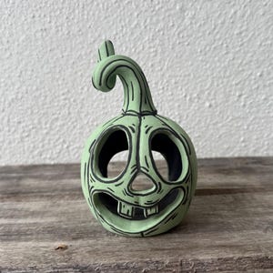 May include: A light green ceramic pumpkin figurine with a carved face featuring large circular eyes, a triangular nose, and a wide mouth with two visible teeth. The pumpkin has a curved stem and black hand-painted outlines.