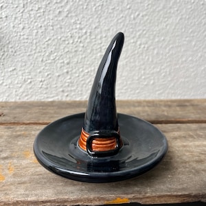 May include: Black ceramic witch hat ring holder. The hat has a tall, curved brim and a wide, flat base. A brown and gold band with a buckle encircles the hat. The ring holder sits on a wooden surface.