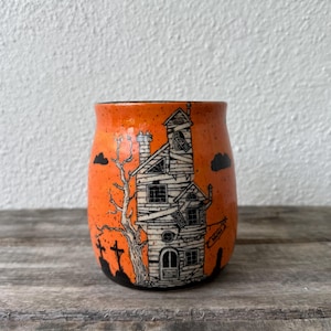 May include: A bright orange ceramic mug with a speckled finish featuring a black ink illustration of a multi-story haunted house, a bare tree, and gravestones. The house design includes boarded windows, a chimney, and a sign hanging from the side.