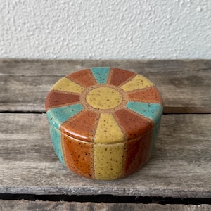 May include: A ceramic trinket box with a lid. The box is round with a colorful design. The lid features a sunburst pattern in yellow, teal, and orange. The sides of the box are striped with the same colors. The box is sitting on a wooden surface.