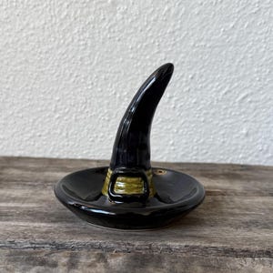 May include: A black ceramic witch hat ring holder with a gold-colored buckle. The hat is glossy and has a curved, pointed top. The base is a shallow dish, perfect for holding rings or small jewelry items. A Halloween-themed decorative item.