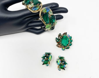 May include: A set of green rhinestone jewelry featuring a bracelet, brooch, and earrings. The jewelry is made of gold-toned metal and features a variety of green rhinestones in different shapes and sizes.