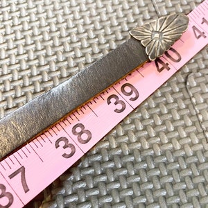May include: A pink measuring tape with black numbers, showing 37, 38, 39, 40, and 41 inches. A silver metal flower-shaped buckle is attached to a dark brown leather strap.