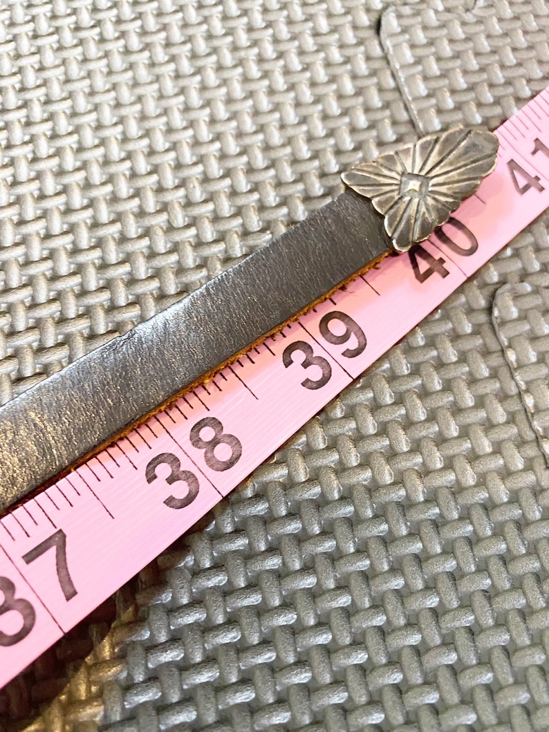 May include: A pink measuring tape with black numbers, showing 37, 38, 39, 40, and 41 inches. A silver metal flower-shaped buckle is attached to a dark brown leather strap.