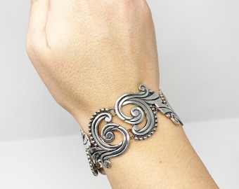 May include: A silver bracelet with a detailed, ornate design featuring swirling patterns and bead accents.