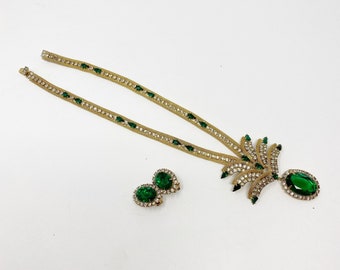 Op de afbeelding: Een gouden ketting met een groene stenen hanger en bijpassende oorbellen. De hanger is een grote, ovale steen omringd door kleinere stenen. De ketting is gemaakt van een ketting met kleine, ronde stenen.