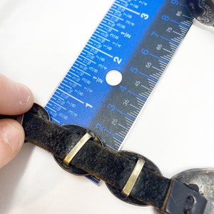 May include: A black leather bracelet with silver-toned metal accents. The bracelet is measured with a ruler, showing it is approximately 2 1/2 inches long.