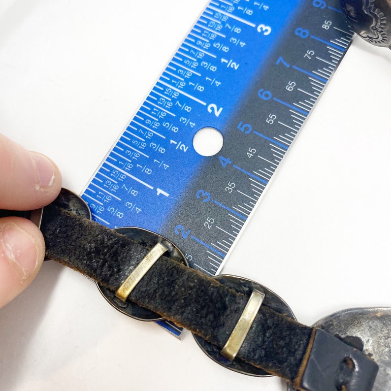 May include: A black leather bracelet with silver-toned metal accents. The bracelet is measured with a ruler, showing it is approximately 2 1/2 inches long.