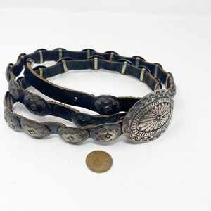 May include: A brown leather belt with silver concho accents. The belt has a large oval concho with a starburst design in the center.
