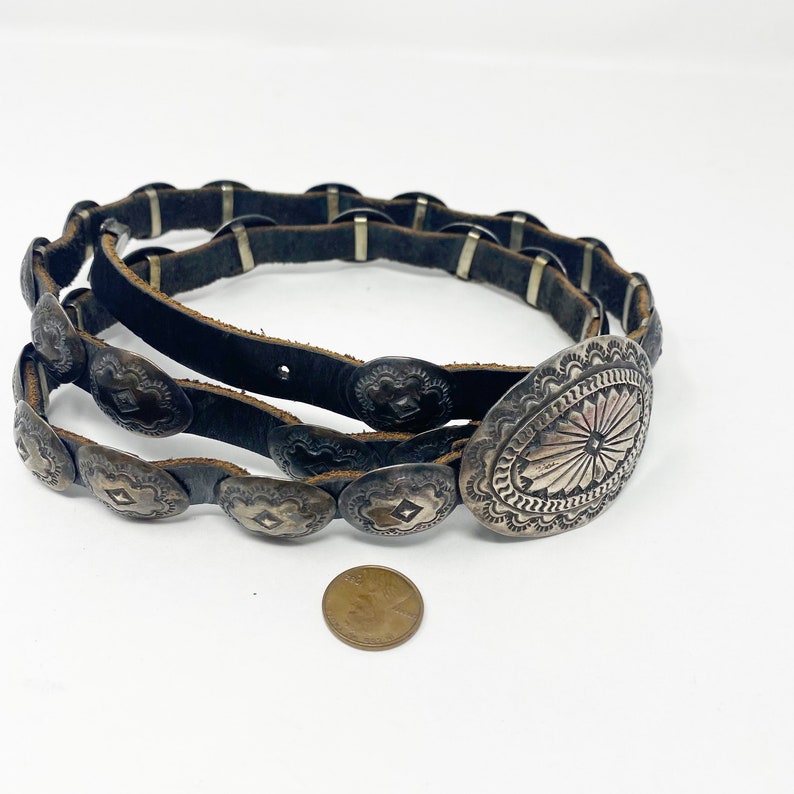 May include: A brown leather belt with silver concho accents. The belt has a large oval concho with a starburst design in the center.