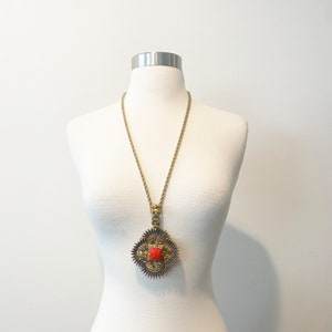 May include: A gold chain necklace with a large, ornate pendant. The pendant is a square shape with a red stone in the center and blue accents around the edges.