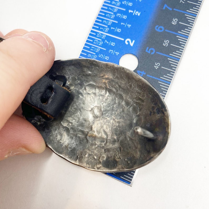 May include: A silver belt buckle with a textured surface and the inscription "STERLING" on the back. The buckle is attached to a black leather strap.
