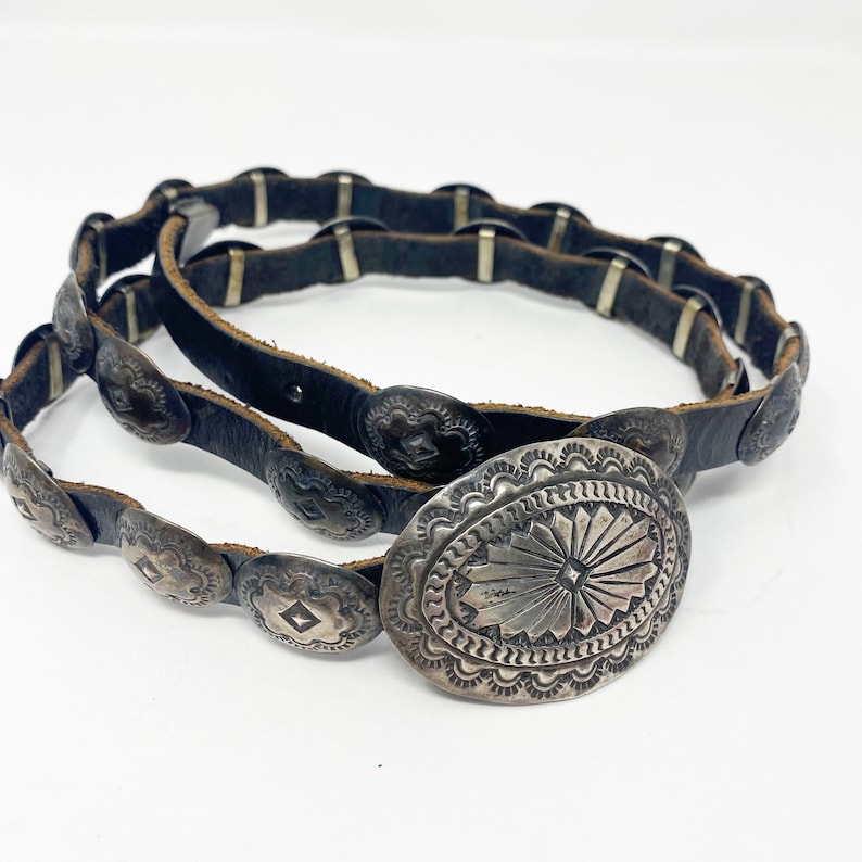 May include: A black leather belt with silver concho accents. The belt has a large oval-shaped buckle with a detailed design.