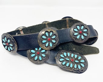 May include: A black leather belt with silver-toned, floral-shaped conchos. Each concho features turquoise-colored stones surrounding a central red stone. The belt is a vintage style, with a classic buckle.