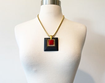 May include: A gold chain necklace with a black and gold square pendant featuring a red square in the center.
