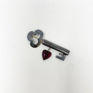 Puede incluir: Una llave de plata con un dije en forma de corazón colgando de ella. El dije está hecho de vidrio rojo.
