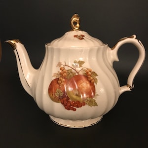 Op de afbeelding: Een witte theepot met een gouden rand en een fruitontwerp. De theepot heeft een deksel met een gouden knop en een gebogen handvat.