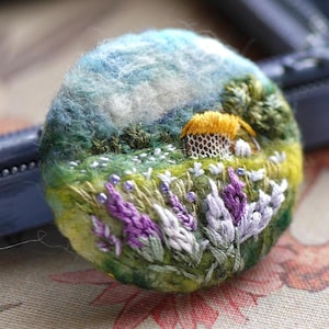 Könnte beinhalten: Eine Filz-Brosche, die eine Landschaftsszene mit einem kleinen Haus, Blumen und einem blauen Himmel darstellt. Die Brosche ist handgefertigt und weist komplizierte Details auf.