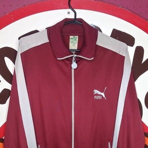 Peut inclure: Veste de survêtement Puma bordeaux avec des rayures blanches sur les manches et les épaules. Logo Puma blanc sur la poitrine et fermeture éclair complète. La veste est suspendue à un cintre noir.