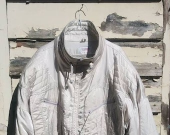 May include: A light beige vintage jacket with a high collar and a zip-up front. The jacket has multiple pockets and a quilted design. It is hanging on a metal hook against a weathered, peeling paint background.