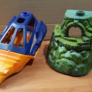 May include: Two vintage toy components. One is a blue and yellow spaceship-like vehicle with a cockpit. The other is a green, rocky structure with an archway. Both are made of plastic and appear to be from a playset.
