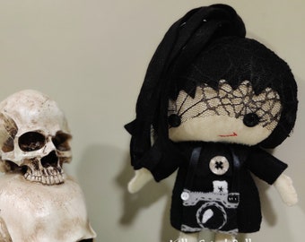 May include: Black and white doll with a black veil and a black dress with a camera design. The doll is standing next to two white skull figurines.