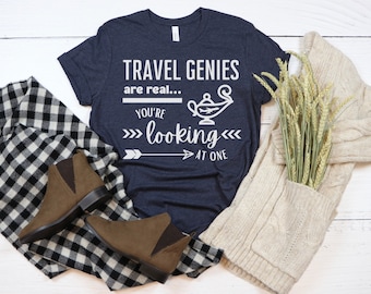 May include: A navy blue t-shirt with the text "TRAVEL GENIES are real... YOU'RE looking AT ONE" and a white graphic of a lamp with a plane inside. The shirt is laid out on a white wooden surface with a brown and white plaid scarf, brown boots, and a white knitted cardigan with wheat stalks.