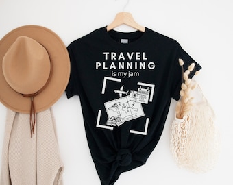 May include: Black t-shirt with the text "TRAVEL PLANNING is my jam" in white. The shirt features a graphic of a world map, camera, and airplane. A tan hat and beige scarf hang to the left.