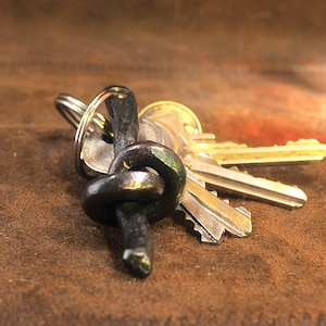 Op de afbeelding: Een handgemaakte, zwarte metalen sleutelhanger met een uniek knoopontwerp. De sleutelhanger bevat meerdere sleutels met zilver- en goudkleurige metalen koppen. Gepresenteerd op een bruin, getextureerd oppervlak.