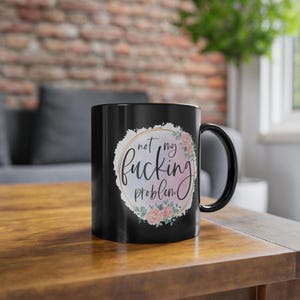 May include: A black ceramic mug featuring a circular design with a watercolor-style pastel background, a gold-colored ring, and pink floral accents. The text "not my fucking problem" is printed in a black cursive script in the center of the design.