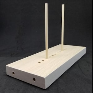 May include: A rectangular light-colored wooden base featuring a series of small circular holes along the top surface and two holes on the front edge. Two thin, vertical wooden dowels are inserted into the top surface of the wooden base.