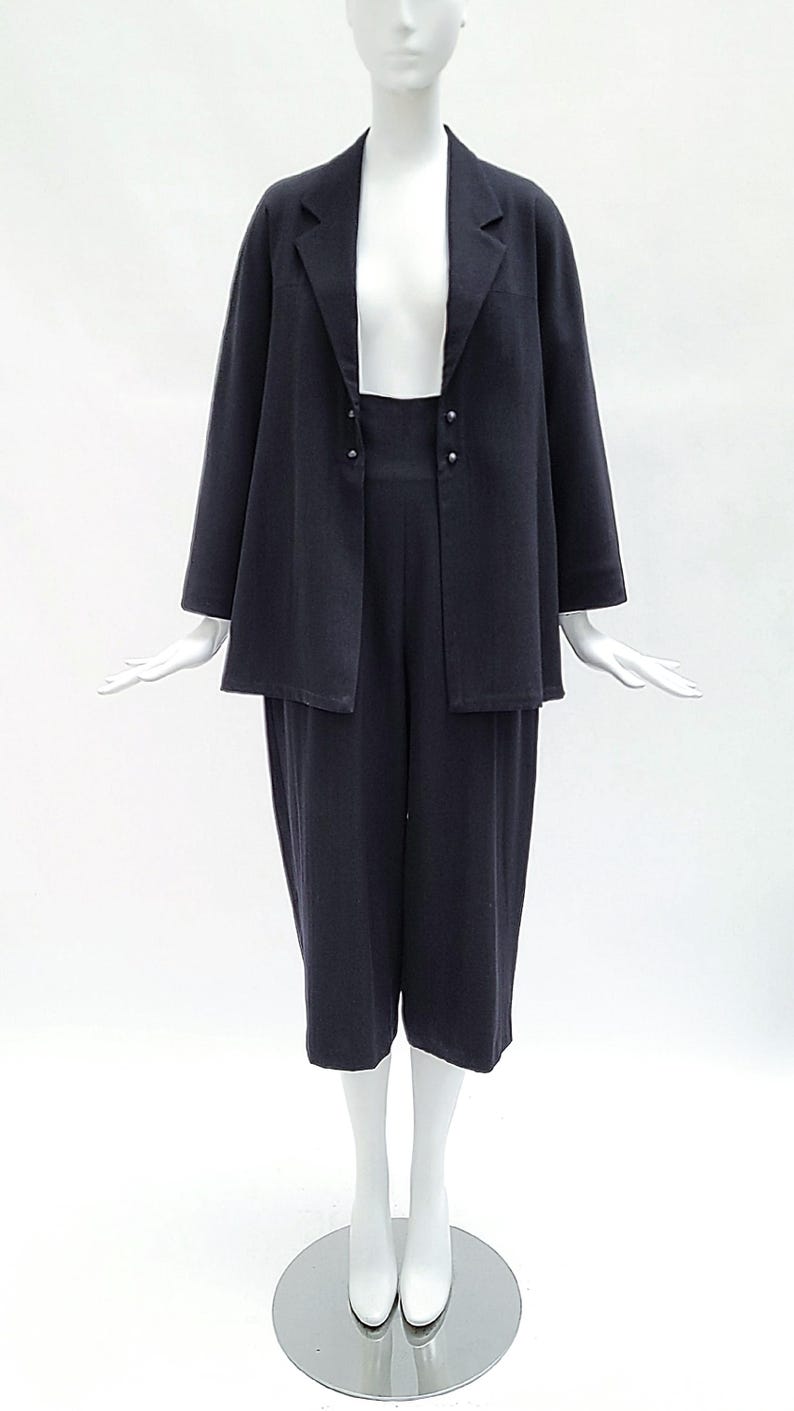 May include: A dark navy blue two-piece set consisting of a long-sleeved blazer with a notched lapel and two small silver-toned buttons, paired with matching high-waisted cropped trousers that feature a wide-leg silhouette and a clean, simple finish.