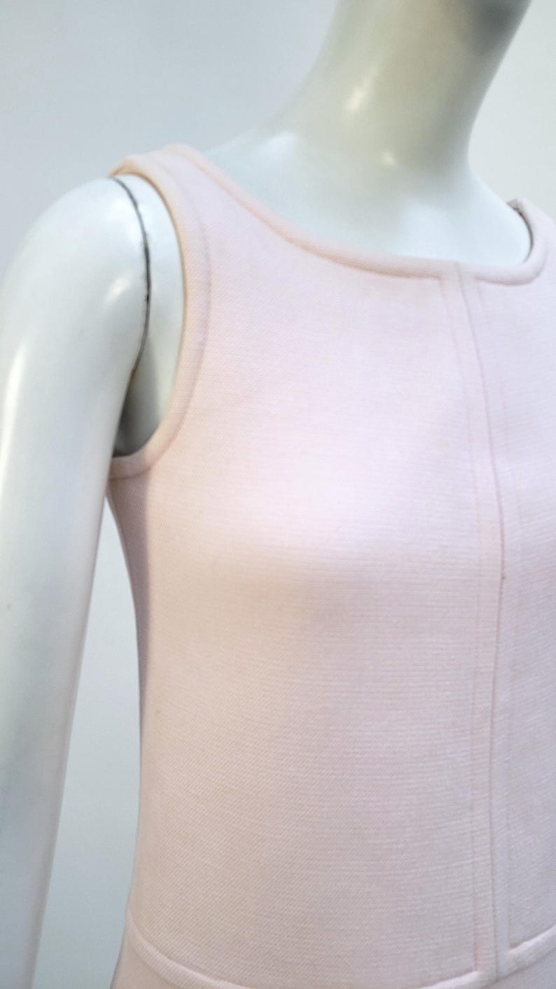 Puede incluir: Un vestido rosa p&aacute;lido sin mangas con cuello redondo y una costura vertical en el centro. El vestido se exhibe en un maniqu&iacute;, destacando su dise&ntilde;o simple y elegante. La tela parece ser un tejido de punto suave.