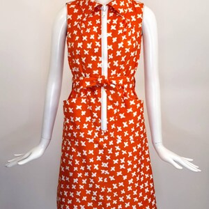 May include: A sleeveless orange dress with a white geometric pattern. The dress has a white zipper down the front and a white belt.