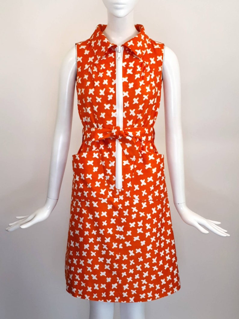May include: A sleeveless orange dress with a white geometric pattern. The dress has a white zipper down the front and a white belt.