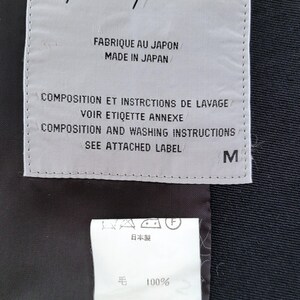 May include: A rectangular white garment label with black text reading Yohji Yamamoto, Made in Japan, and size M, positioned above a smaller white care tag with Japanese text, 100% wool content, and laundry symbols on a black fabric background.