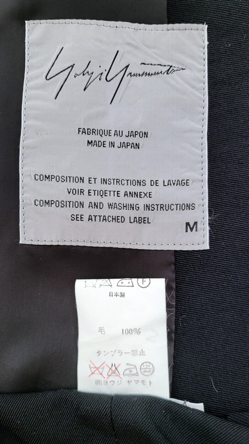 May include: A rectangular white garment label with black text reading Yohji Yamamoto, Made in Japan, and size M, positioned above a smaller white care tag with Japanese text, 100% wool content, and laundry symbols on a black fabric background.