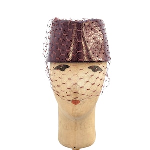 May include: A cylindrical pillbox hat in a deep metallic burgundy with a textured, reptilian-like finish. The hat is adorned with a matching burgundy netting veil that drapes over the front, creating a structured and elegant appearance.