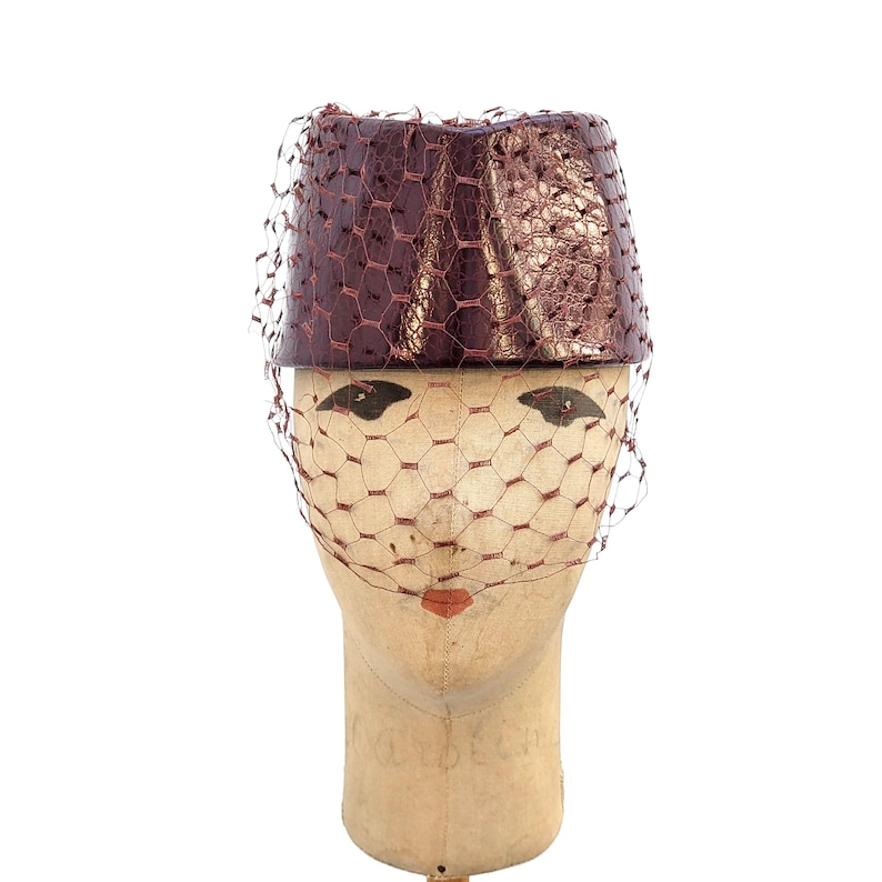 May include: A cylindrical pillbox hat in a deep metallic burgundy with a textured, reptilian-like finish. The hat is adorned with a matching burgundy netting veil that drapes over the front, creating a structured and elegant appearance.